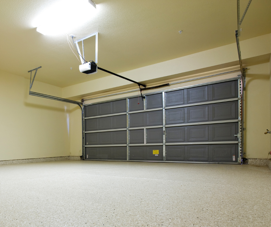 interior view of a clean garage in San Diego, California with a closed, sectional, roll-up garage door. Above the door is a garage door opener unit with its track leading towards the back of the garage, and the floor is finished with a speckled epoxy coating. The walls are painted in a light color, and the ceiling has a centrally mounted light fixture, illuminating the space.